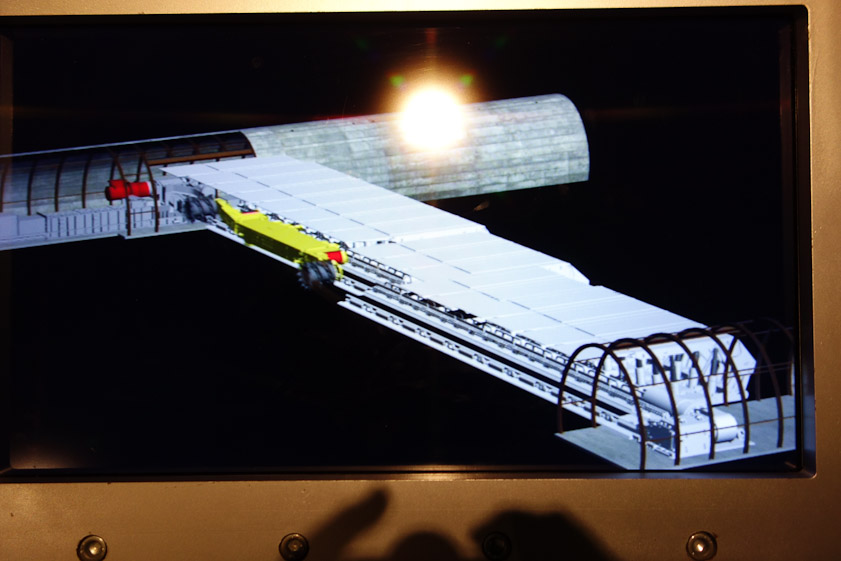 This snapshot of a video shows a seam of coal being dug out between two horizontal tunnels. The yellow machine is called a coal shearer or coal plough, and moves back and forth, widening the seam as the white units hold up the ceiling.