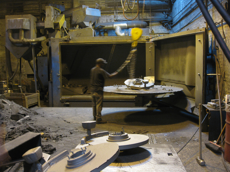 After the molds have cooled, they are taken apart, the casting moved to this turntable, and the door on the right swings closed, taking the casting into the chamber beyond the man, where it is blasted with stell pellets to clean it.