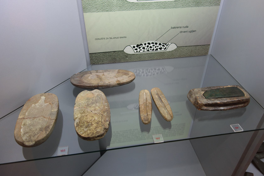 Molds for casting metal objects. An axe head is in its mold at the right. In the back is a diagram of a copper smelting charcoal fire.