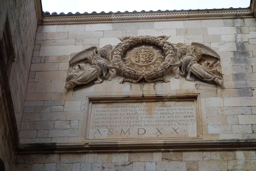 Twwo stone angels support the monogram of Christ over the Sponza courtyard. The date is 1520.