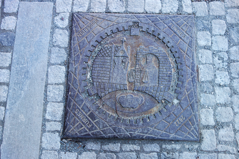 Manhole cover in Trondheim. A crowned figure on the right holding scales of justice symbolizes the secular rulers. The figure on the left with a crozier and making a blessing gesture symbolizes eccesiastical powers. Three faces at the bottom may be the untitled lay population.