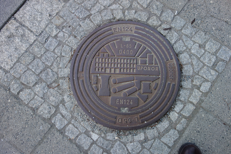 Manhole cover in Bodø. There is a Finnish company UPONOR that makes among other things manhole covers. This one shows a factory above and perhaps a forge and rolling mill below. The symbology and relation to Bodø are unexplained.