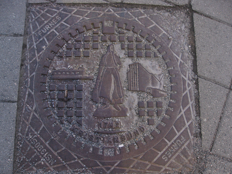 Manhole cover in Kristiansund. The central figure is the Klippfiskkjerringa statue of the wife of a klippfisk worker. The boat to the left may be one of the several ferries joining the four islands of Kristinasund. The building on the right is Kirkelandet church, built in 1964 by the architect Odd Østbye.