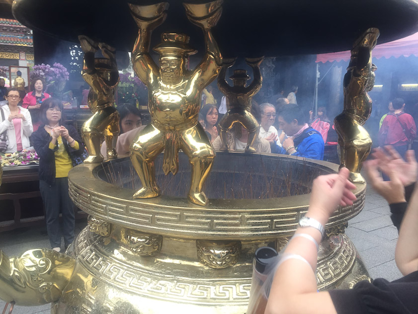 Brass strongmen holding up the cover of the brazier as people wave the clouds of incense smoke over themselves.