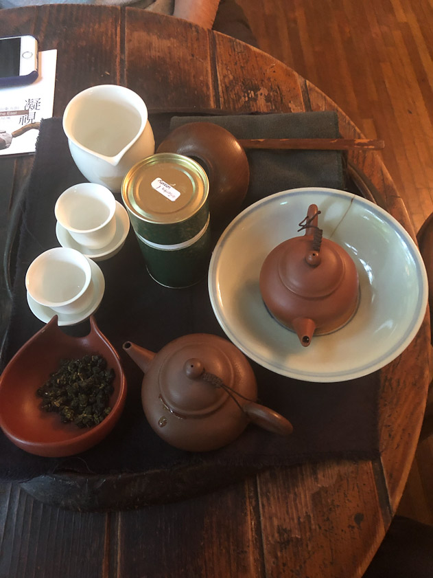 The gear for tasting tea: a scoop holding the first tea, a cannister holding the second tea, two teapots in which to brew the two teas, a pitccher into which the tea pot is emptied, and tea cups filled from the pitccher.