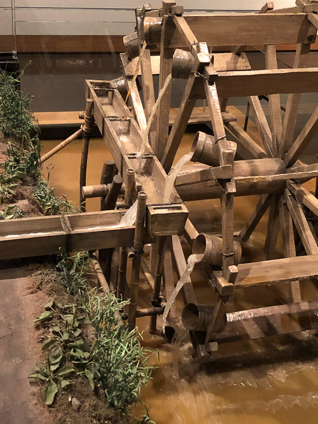 The wheel is driven by the river current, the buckets fill when they are under water, and it begins to spill out as the buckets emerge. Some of their contents are captured in the trough.