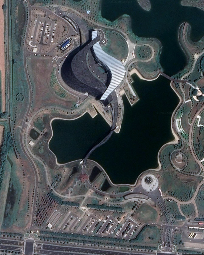 This satellite photo shows the southern entrance at the bottom, the curved pedestrian bridge corssing the lake, and the two curving, intersecting buildings of the museum, black on the left and white on the right. The entrance is in the white building near where it crosses over the black building.