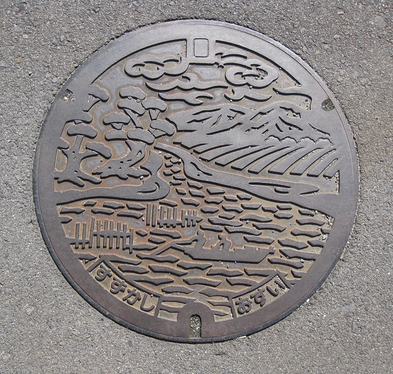 Manhole cover in Suzuka. The kana on the left read suzukashi (Suzuka City) and those on the right read osui (sewage). The design above shows fishermen with rows of tea bushes on the shore on the right and pine trees on the left.