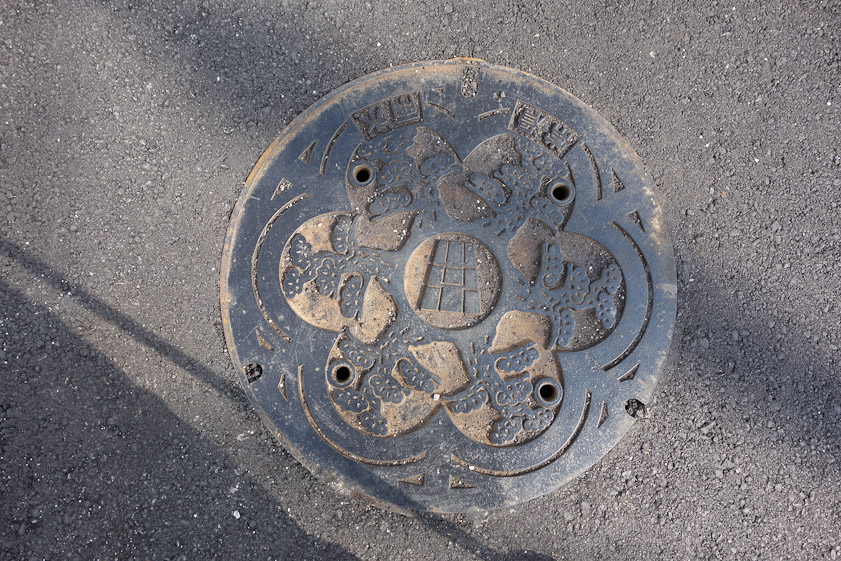 Manhole cover in Tokoname on 16 May 2013. The shape in the center probably represents one of the many tall, brick chimneys of the oil- and coal-fired kilns in Tokoname that used to fill the air with smoke. The six lobes surrounding it may be clouds of smoke.