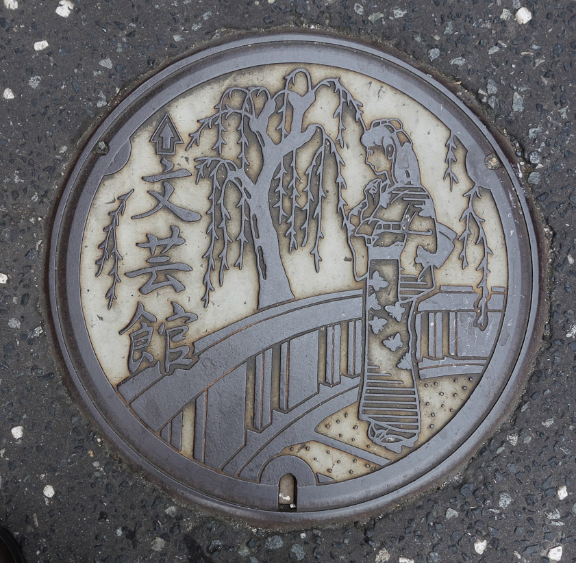 Manhole cover in Kinosakionsen on 27 May 2013. Visitors go from bath to bath wearing yukatas and wooden clogs like those she wears.