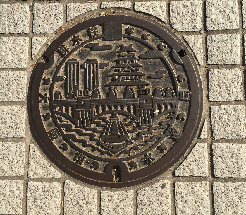 Manhole cover in Osaka. Osaka Castle on the rigt, skycrapers on the left, the Suishou Bridge in Nakanoshima, and a tourist boat cruising under it.