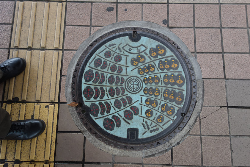 Manhole cover in Akita. The meaning of these patterns was revealed when we visited the Kanto festival hall. Shown here are two towers of lanterns carried in parades during the Kanto matsuri.