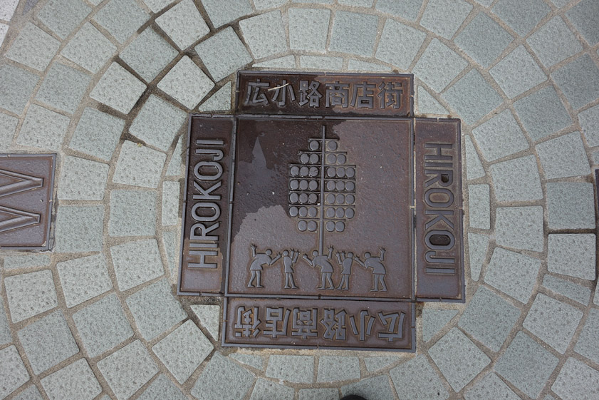 Street marker in Akita showing a man balancing a tall array of lanterns on his shoulder during the Kanto matsuri parade. Hirokoji is a district.