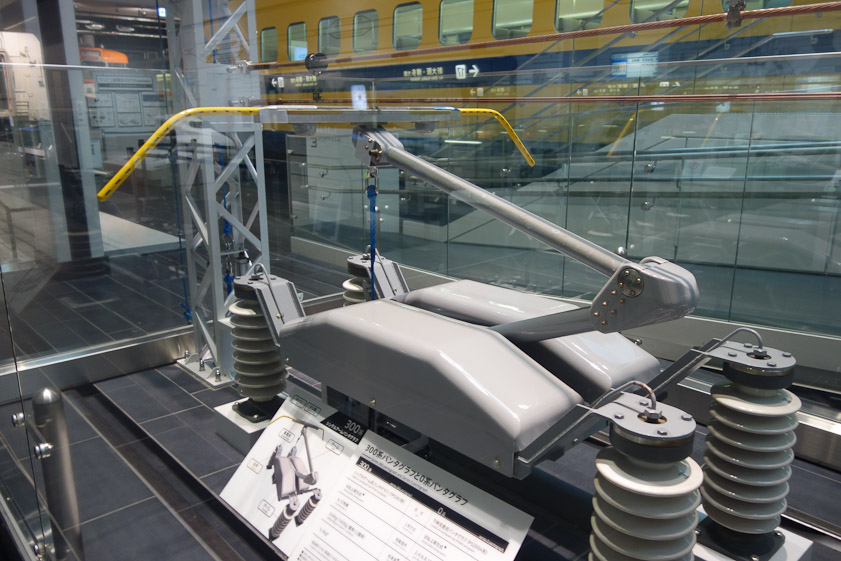 The yellow pantograph mounted on top of a shinkansen engine is in continuous contact with the trolley wire, drawing current to power the train.