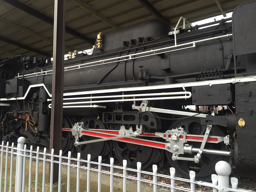 The silver rod just above the knob on the fence at lower right is the piston rod. It drives the weels (four on this side) via the red beams. The smaller silver rods operate the valve that controls the entry of hot steam to, and the exhaust of expanded steam from, the cylinder.