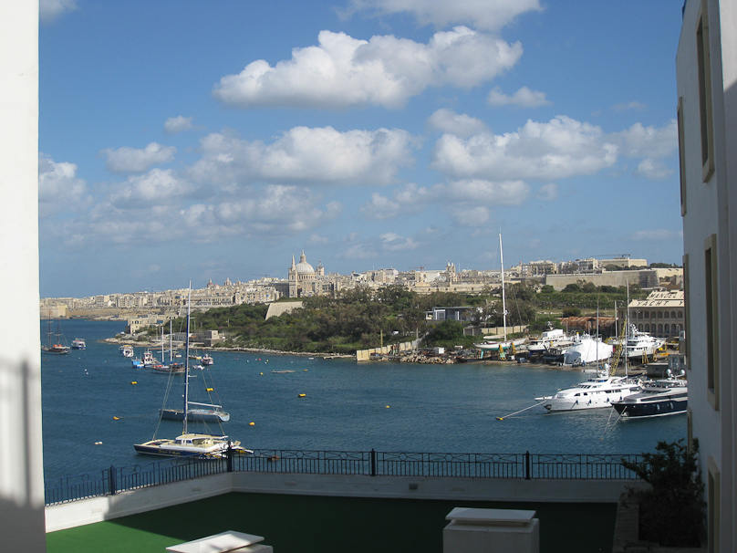 View out our hotel window in Sliema, adjacent to Manoel Island (trees and boat yards) with Valletta visible beyond (dome of the cathedral, wall of fortifications to the right of the tallest mast).
