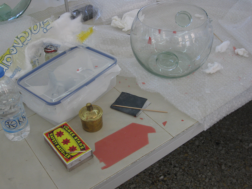 Pieces of glass are temporarily held in place with red wax that is heated by the small spirit lamp to make it adhesive. When the pieces are closely fit, permanent liquid adhesive can be applied and flows into the very narrow channel between pieces by capillary action. The wax patch is then heated and removed.