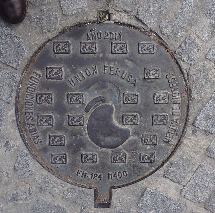 Manhole cover in Segovia. This was made in 2011. Fundiciones Arias is a foundry in Medina de Rioseco in the province of Valladolid. The little squares show two men pouring molten metal in a foundry. Union Fenosa was a gas company, since acquired by Gas Natural.