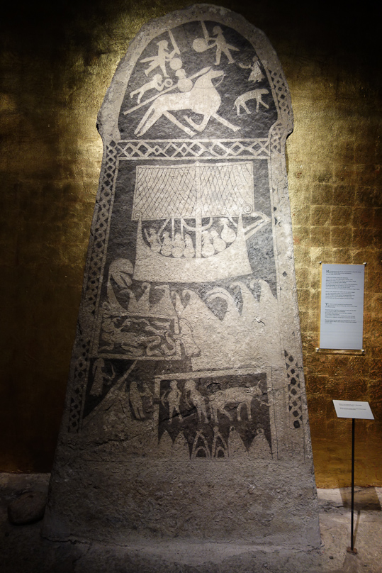 Label: A good example of the picture stones from the late Iron Age, the Viking Age... might be a memorial to a man, who has died a hero. Borne by the ship of death, he is going to Valhalla.. There, a woman, perhaps a Valkyrie, welcomes him, handing him a horn of mead. The scene below the si may illustrate a saga in which Gunnar is thrown into a snakepit to die.